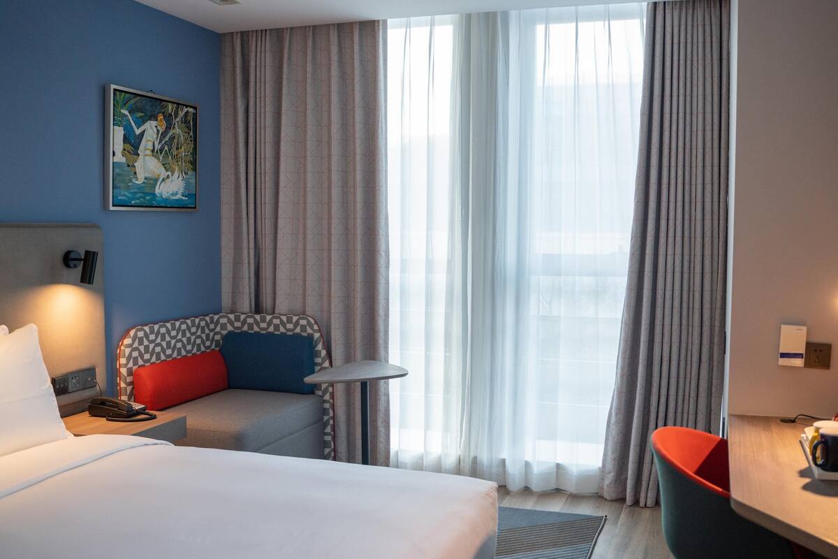 A hotel room with a bed, a desk with a chair, a small table, a telephone, a painting on the wall, and large windows with curtains.