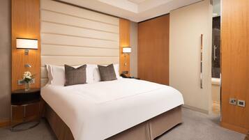 A hotel room with a large bed, two bedside tables, a nightstand, a lamp, a vase with flowers, and a view of a tiled wall.