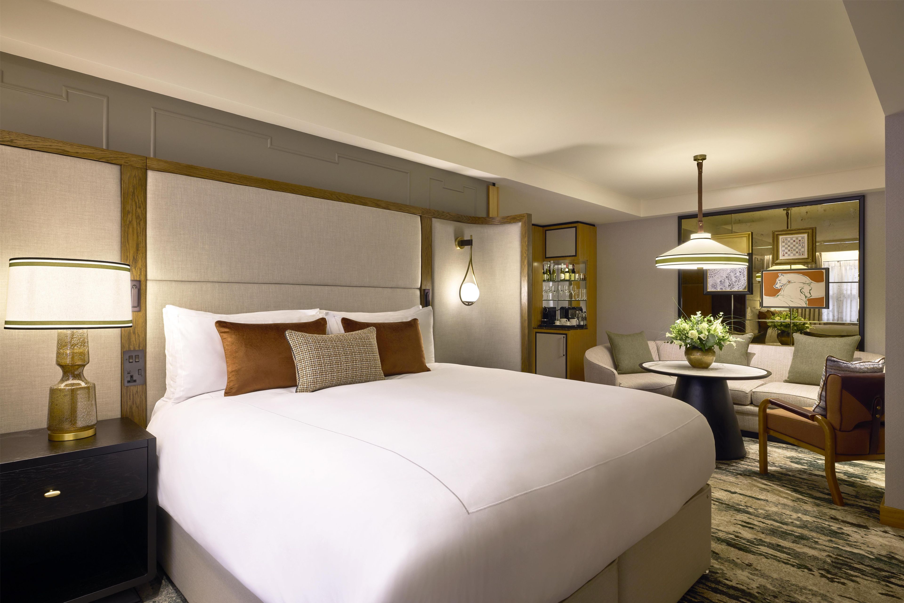 A hotel room with a large bed, bedside table, lamp, chair, round table, and a mirror.