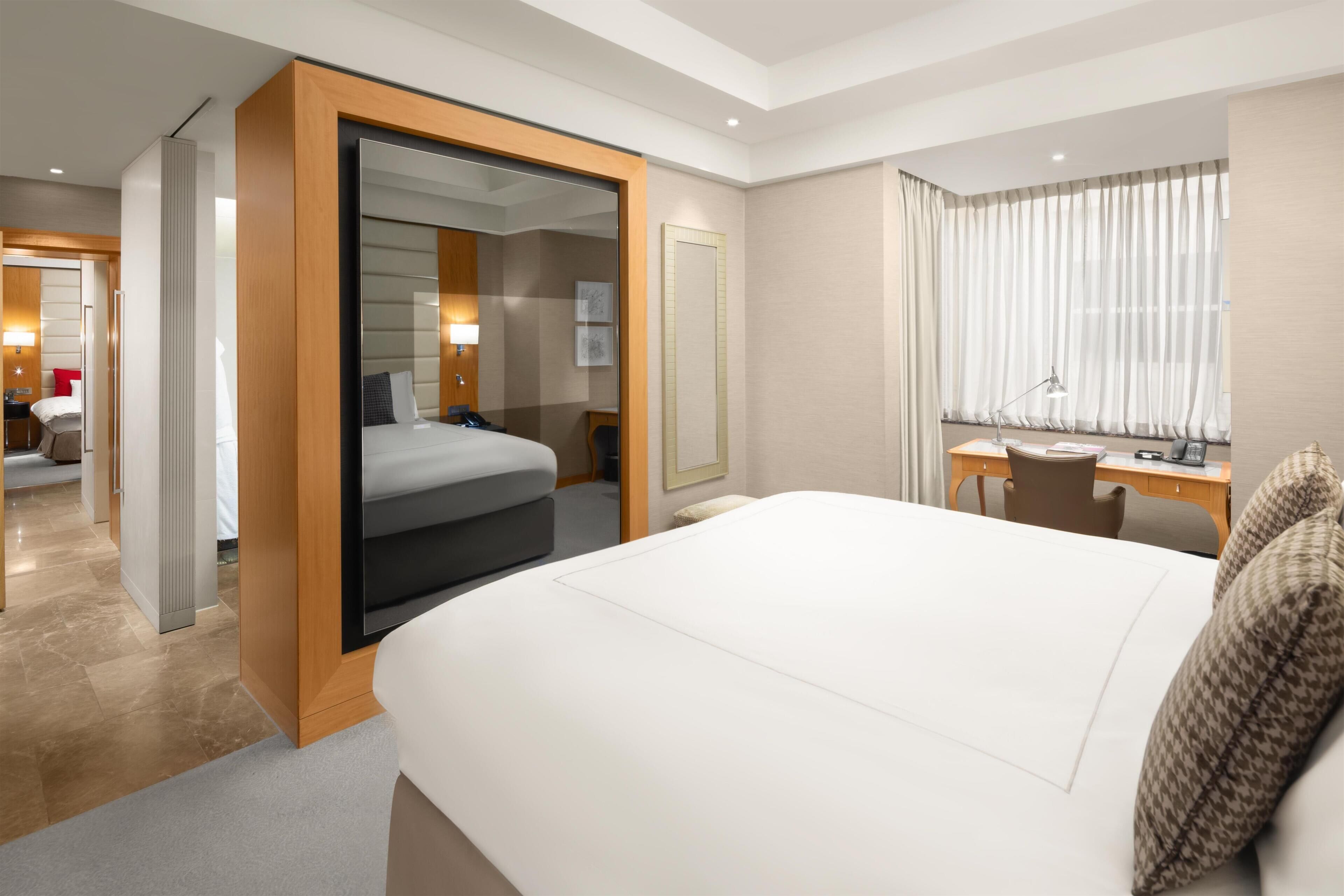 A hotel room with a large bed, a desk, a chair, a mirror, and a door leading to another room.