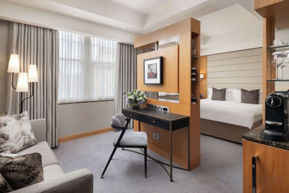 A hotel room with a bed, a desk, a chair, a sofa, and a large window with curtains.