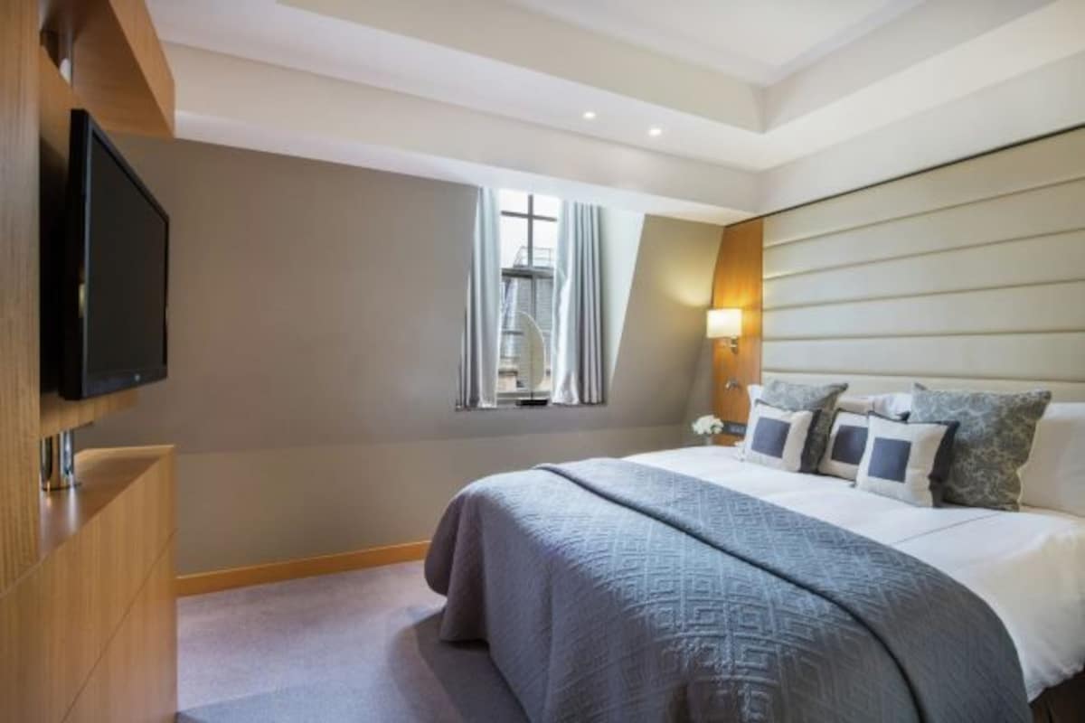 A modern hotel room with a large bed, a flat-screen TV mounted on a wooden cabinet, a window with curtains, and a wall-mounted lamp.