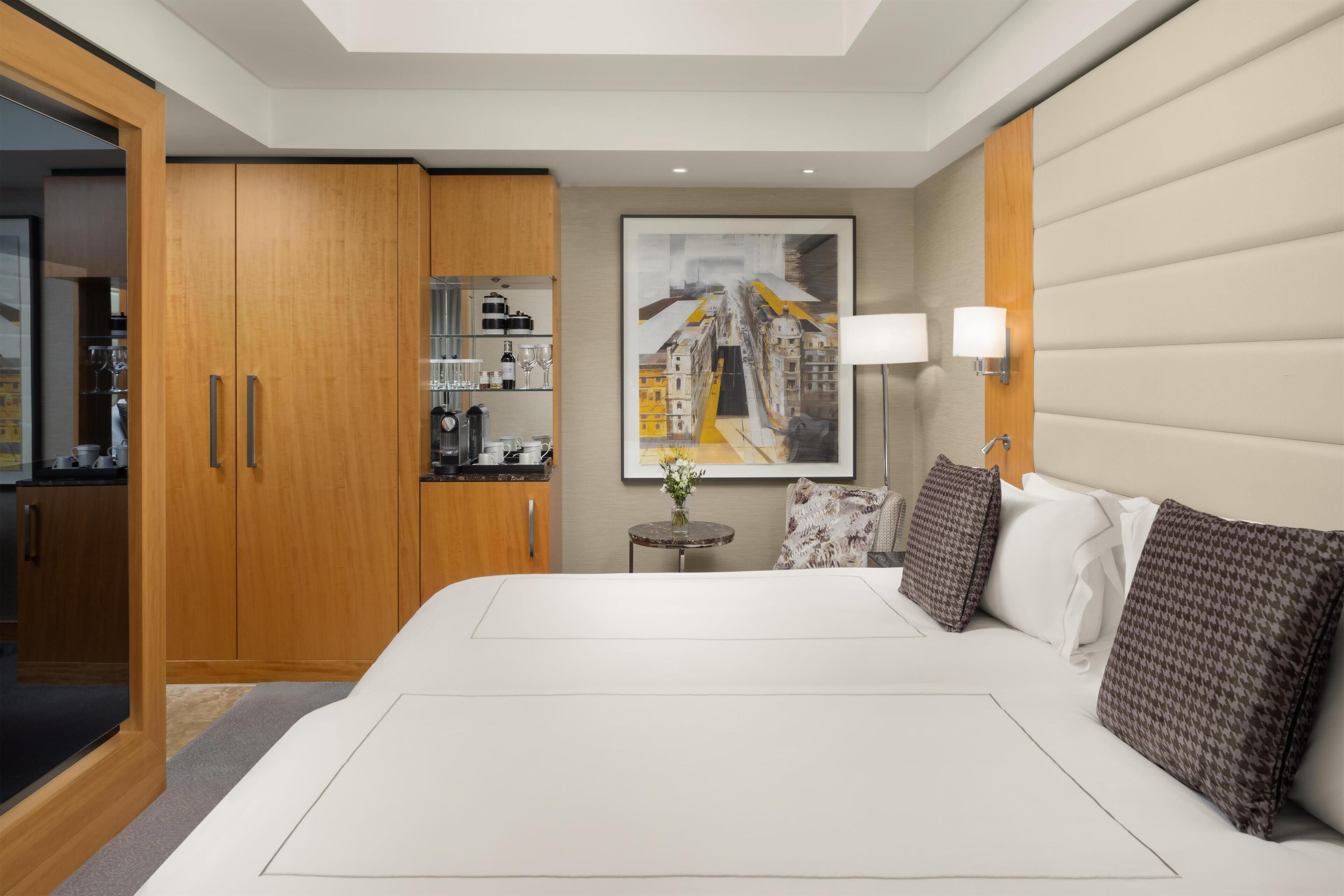 Una habitación de hotel moderna con una cama grande, un armario de madera, una pequeña mesa redonda con un florero y un cuadro de un paisaje urbano en la pared.
