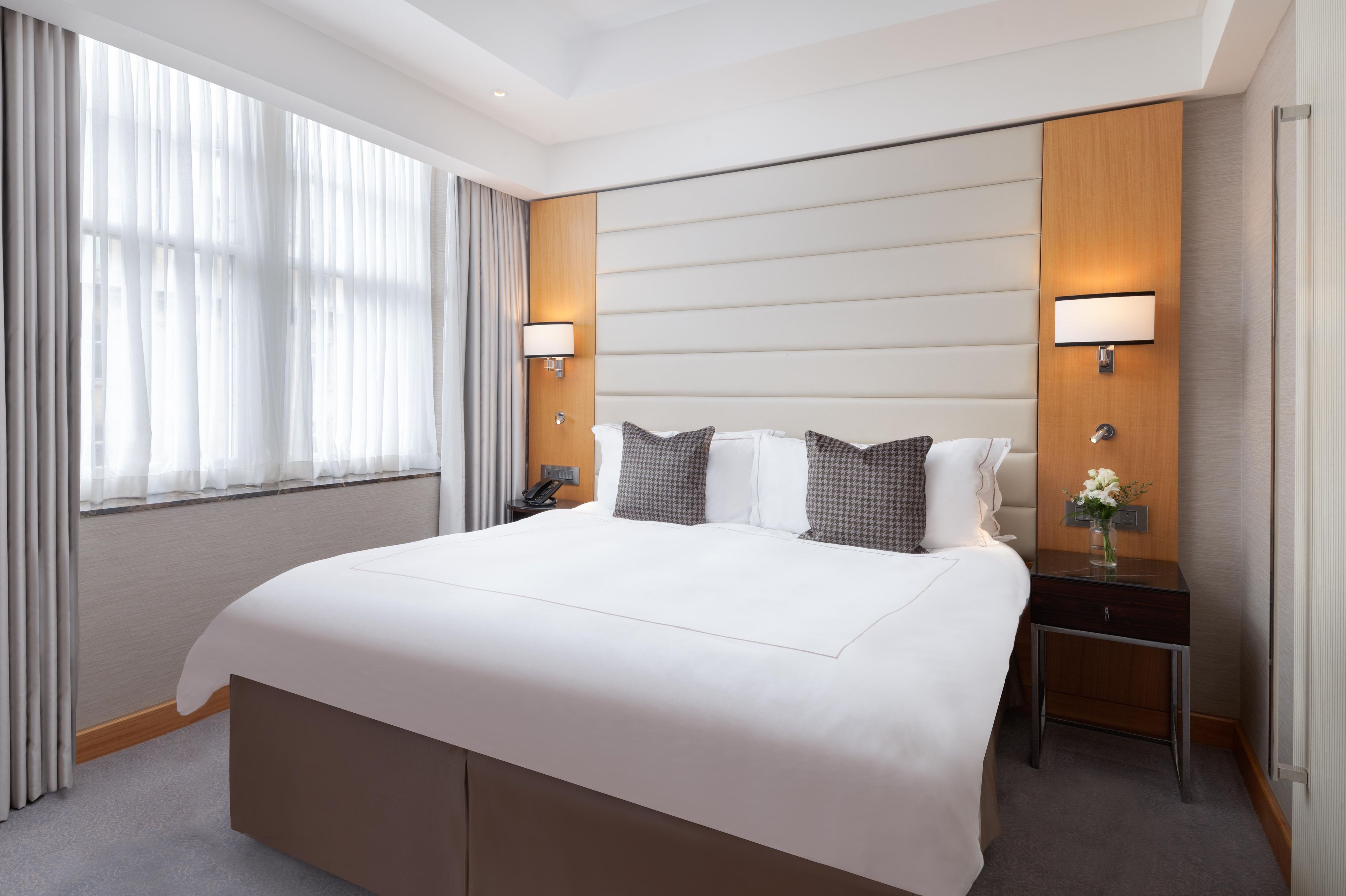 A hotel room with a large bed, two bedside tables, a nightstand, a lamp, and a window with curtains.