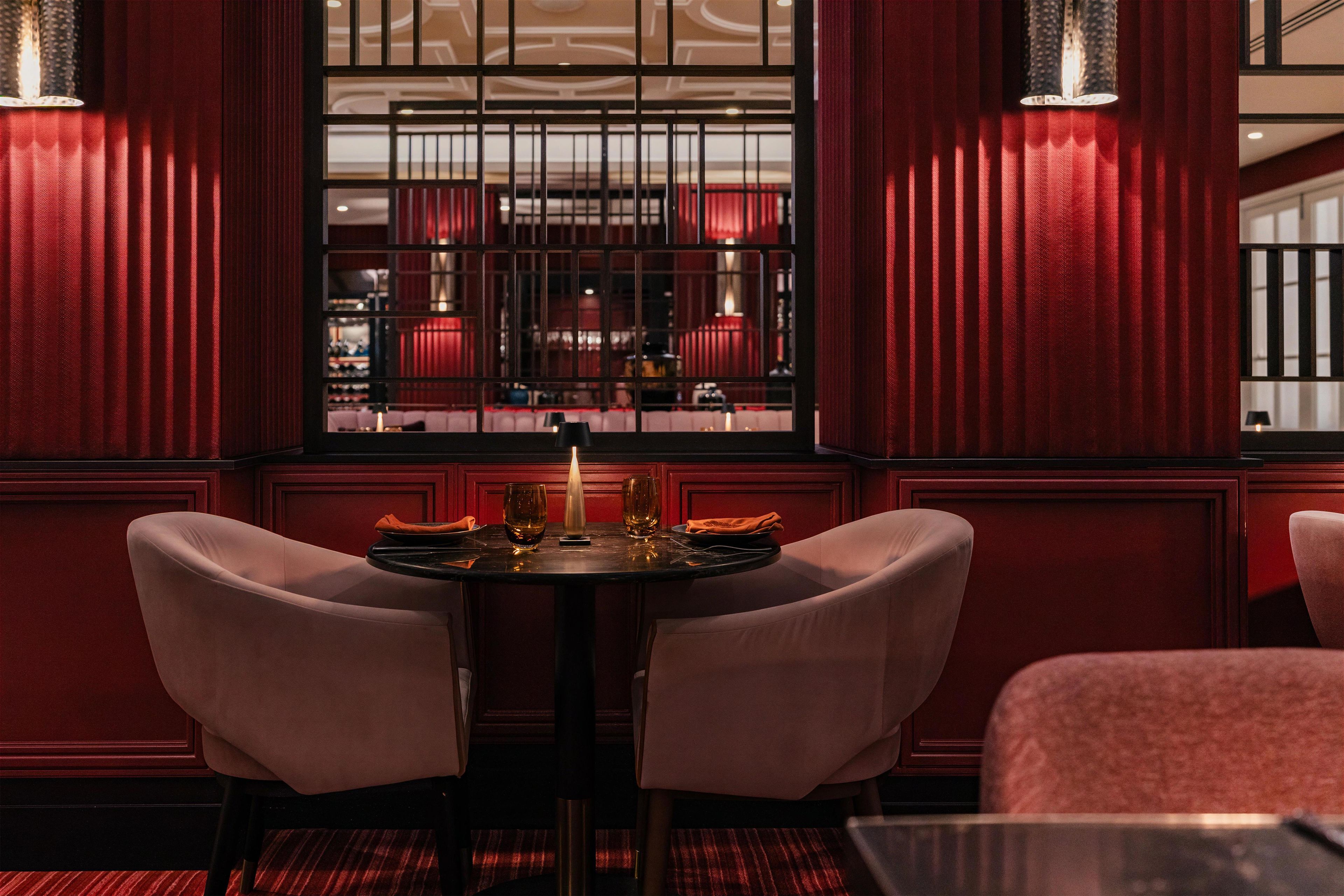 A dining area with a table and chairs, a window with a view of a bar area, and red wall paneling.