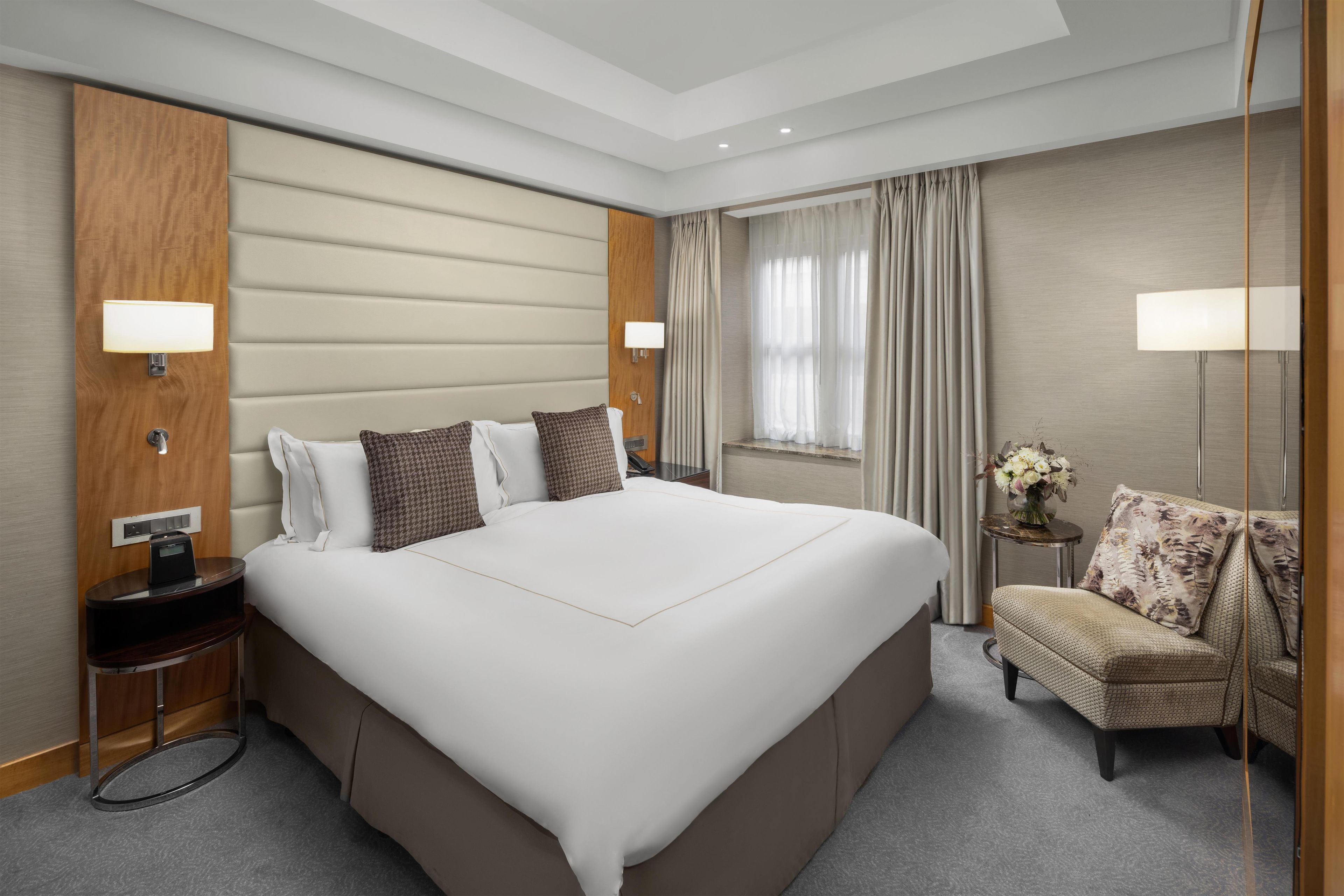 A hotel room with a large bed, bedside lamps, a small table with a vase, and a chair.