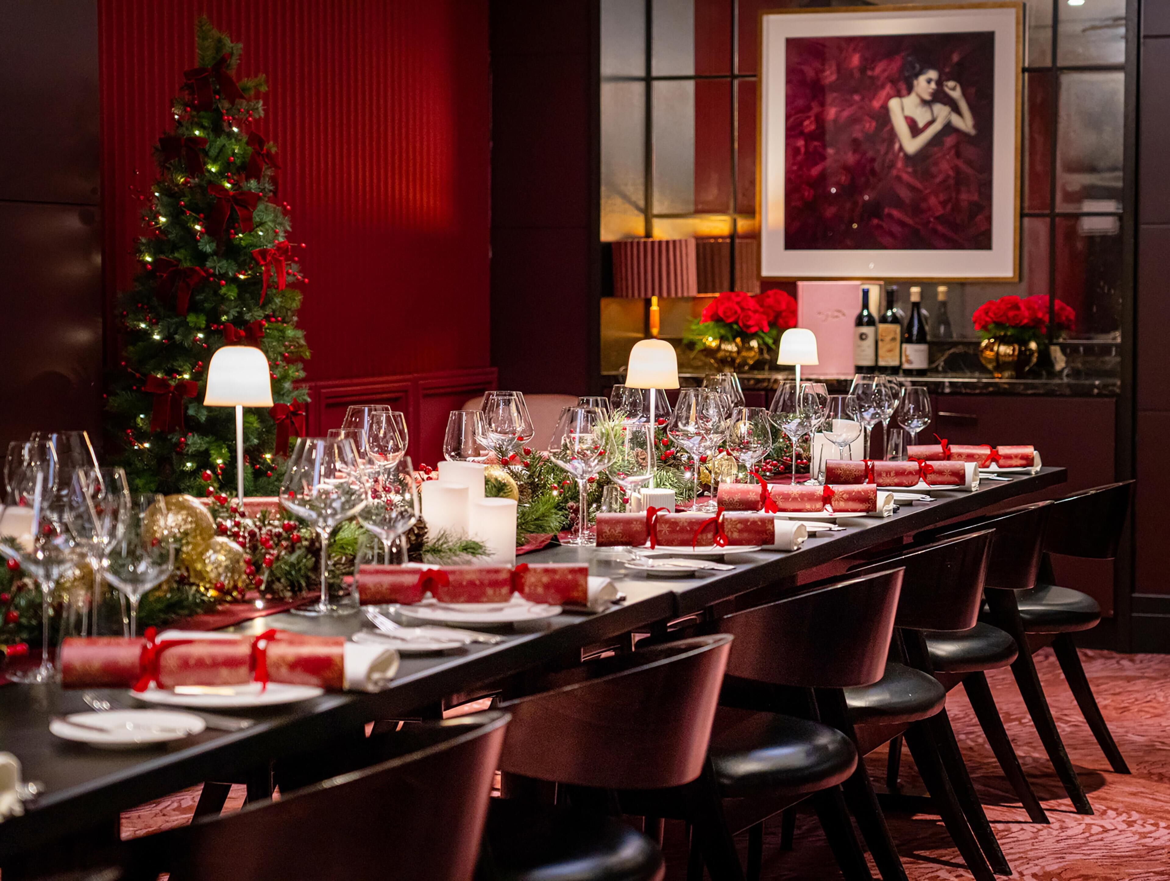 A long dining table set with multiple wine glasses, candles, and red napkins, with a Christmas tree and red decor in the background.