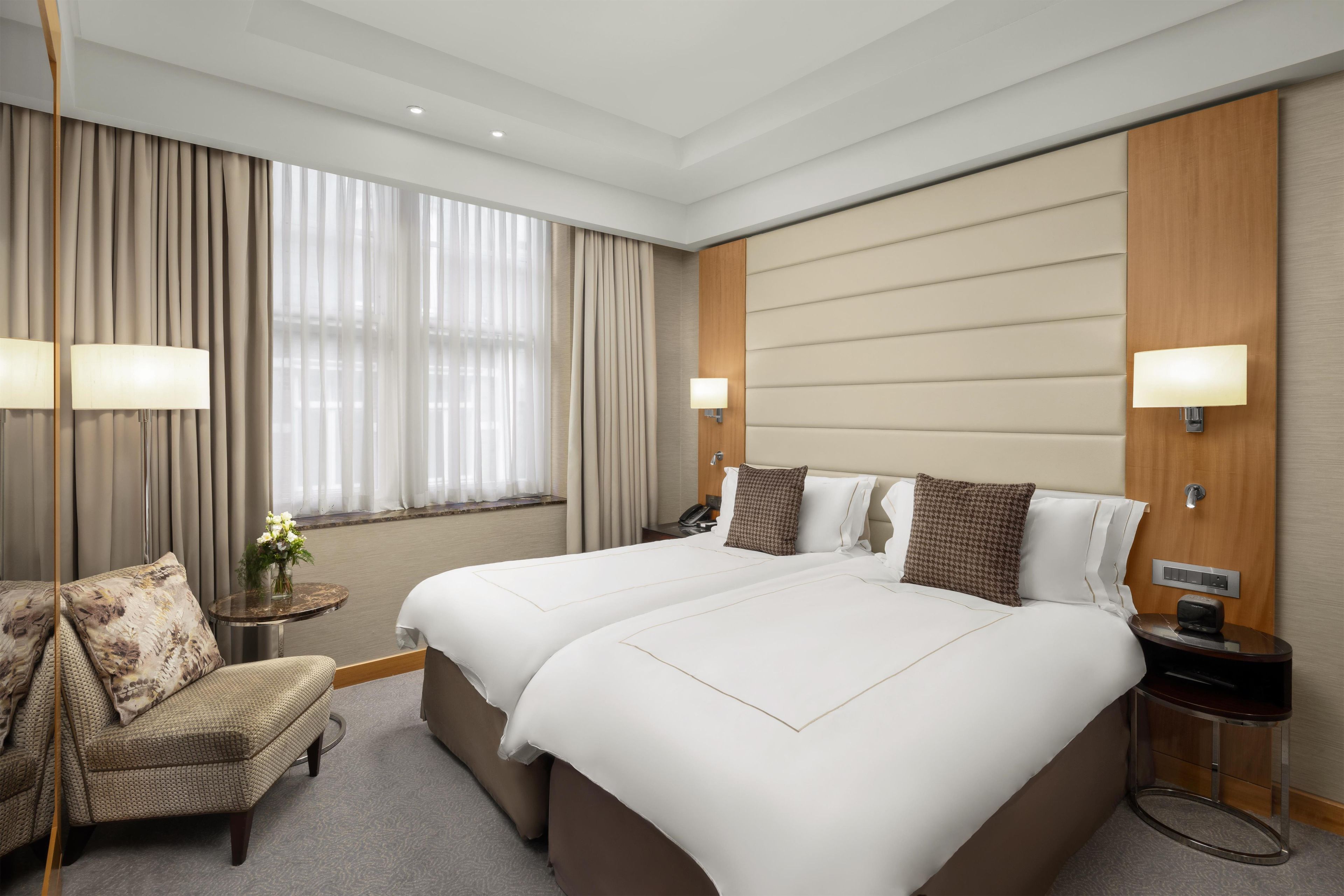 Habitación de hotel con dos camas, una zona de estar con una silla, una mesita con un jarrón de flores y una mesita de noche con lámpara y teléfono.