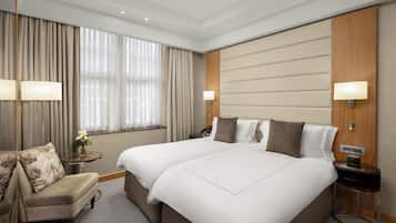 Habitación de hotel con dos camas, una zona de estar con una silla, una mesita con un jarrón de flores y una mesita de noche con lámpara y teléfono.