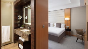 A hotel room with a marble bathroom vanity, a bed with white linens, a bedside table with a lamp, and a chair. - Conrad London St. James (London)