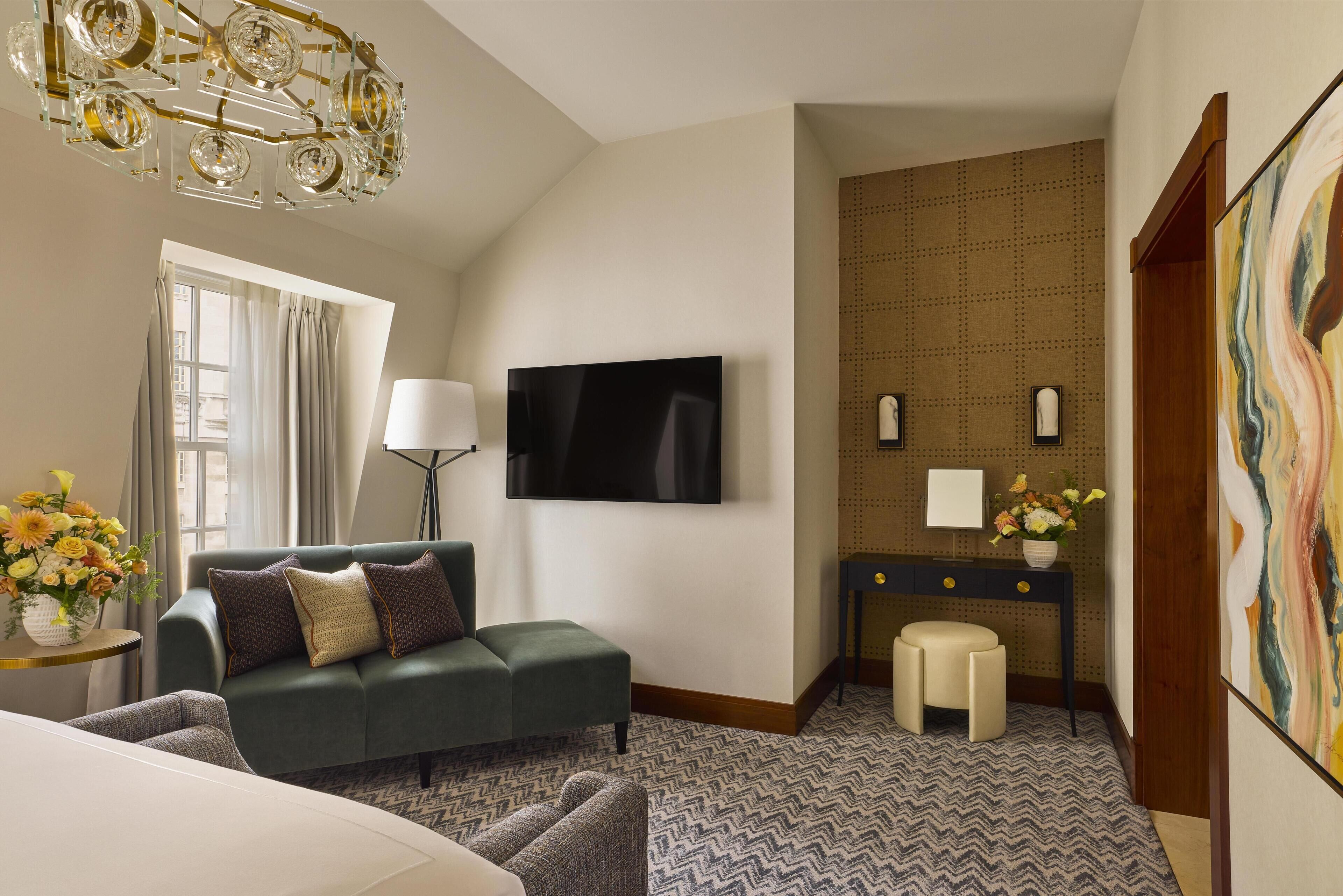 A hotel room with a green sofa, a flat-screen TV, a desk with a lamp, a patterned carpet, and a large abstract painting.