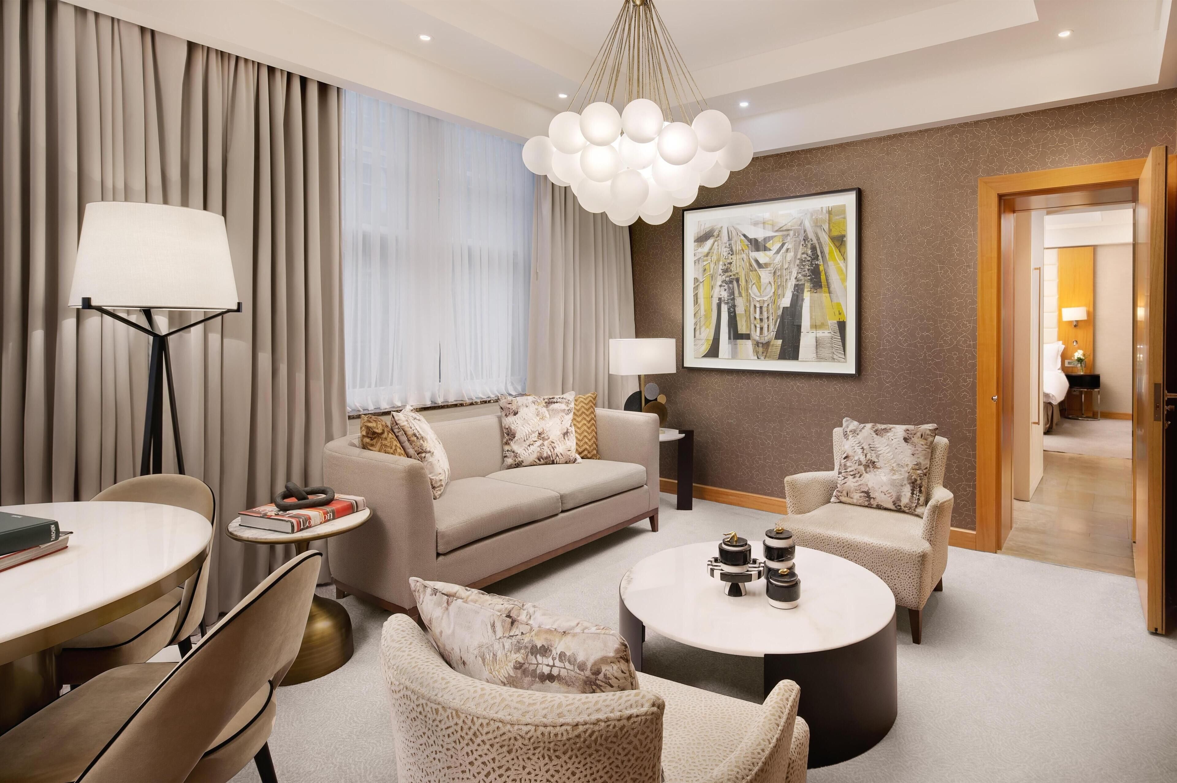 Ett modernt hotellrum med en soffa, fåtöljer, ett runt soffbord, ett sidobord med böcker, en golvlampa, en tavla och en dörr som leder till ett annat rum.