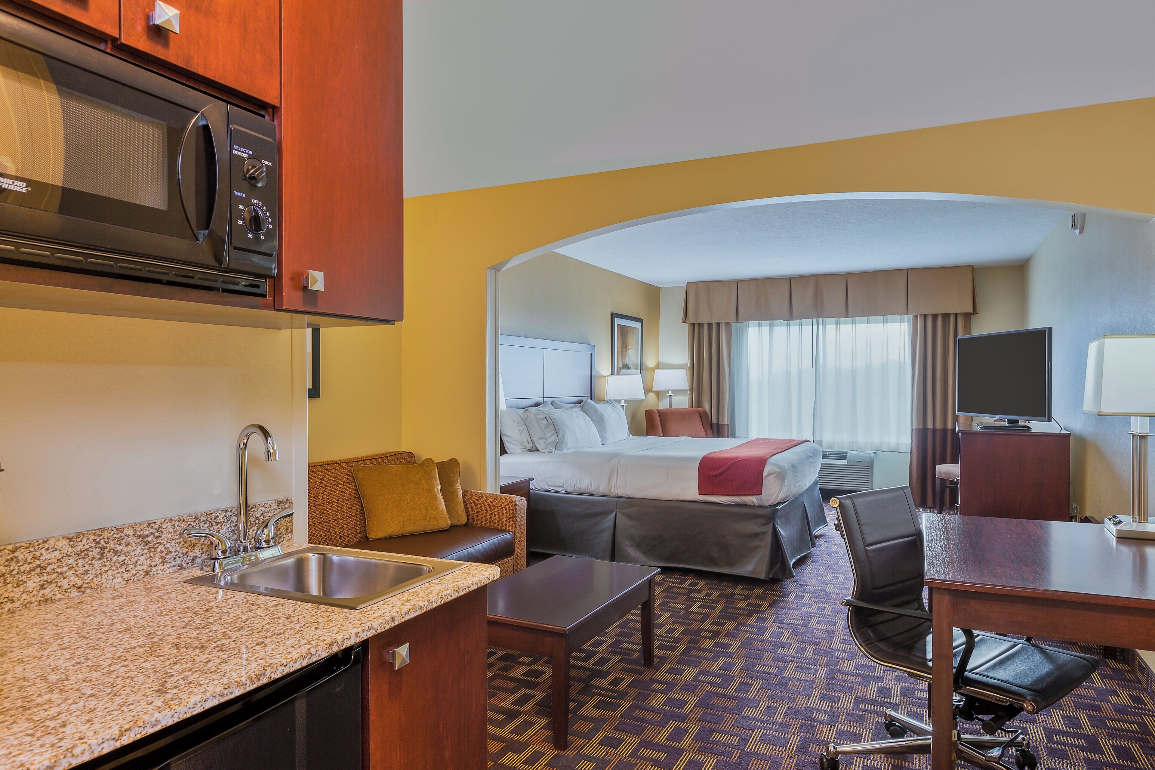 A hotel room with a kitchenette, a bed, a desk, and a TV.