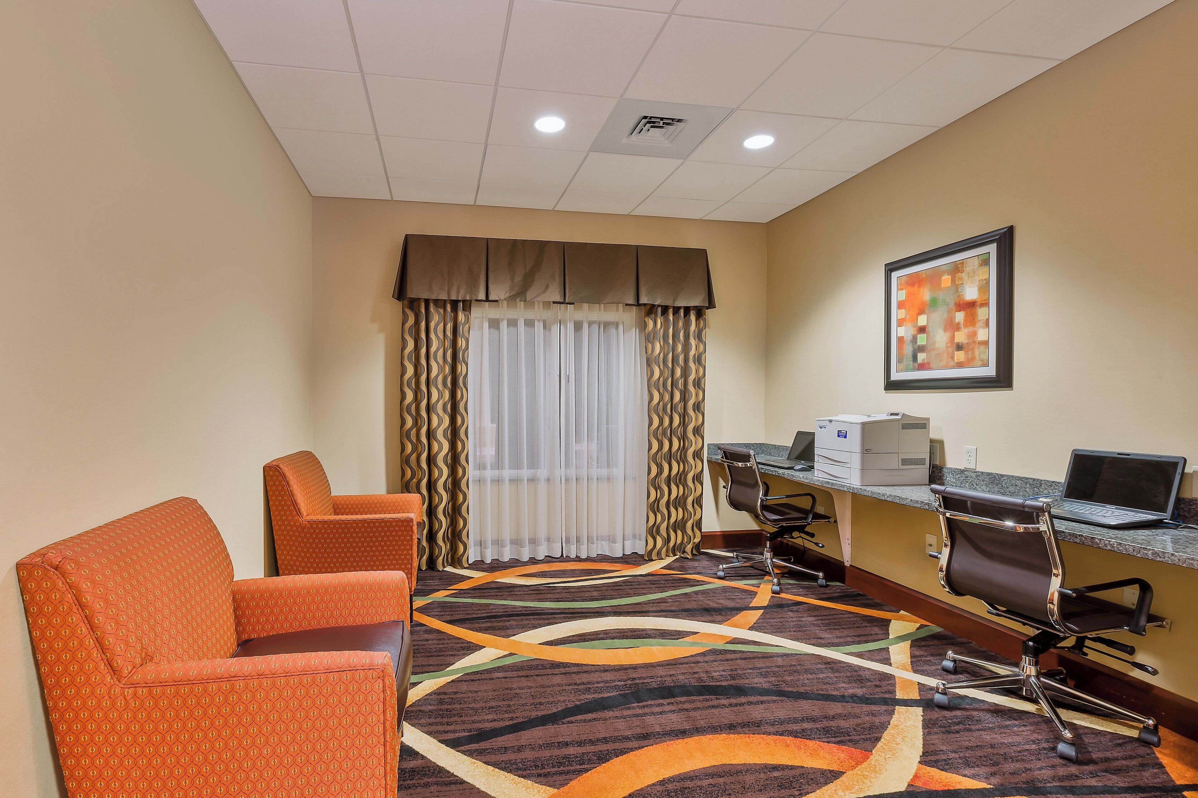 A hotel room with a desk, two chairs, a patterned carpet, a window with curtains, and a framed picture on the wall.