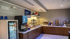 A modern breakfast area with a refrigerator, coffee machines, and a variety of food items.