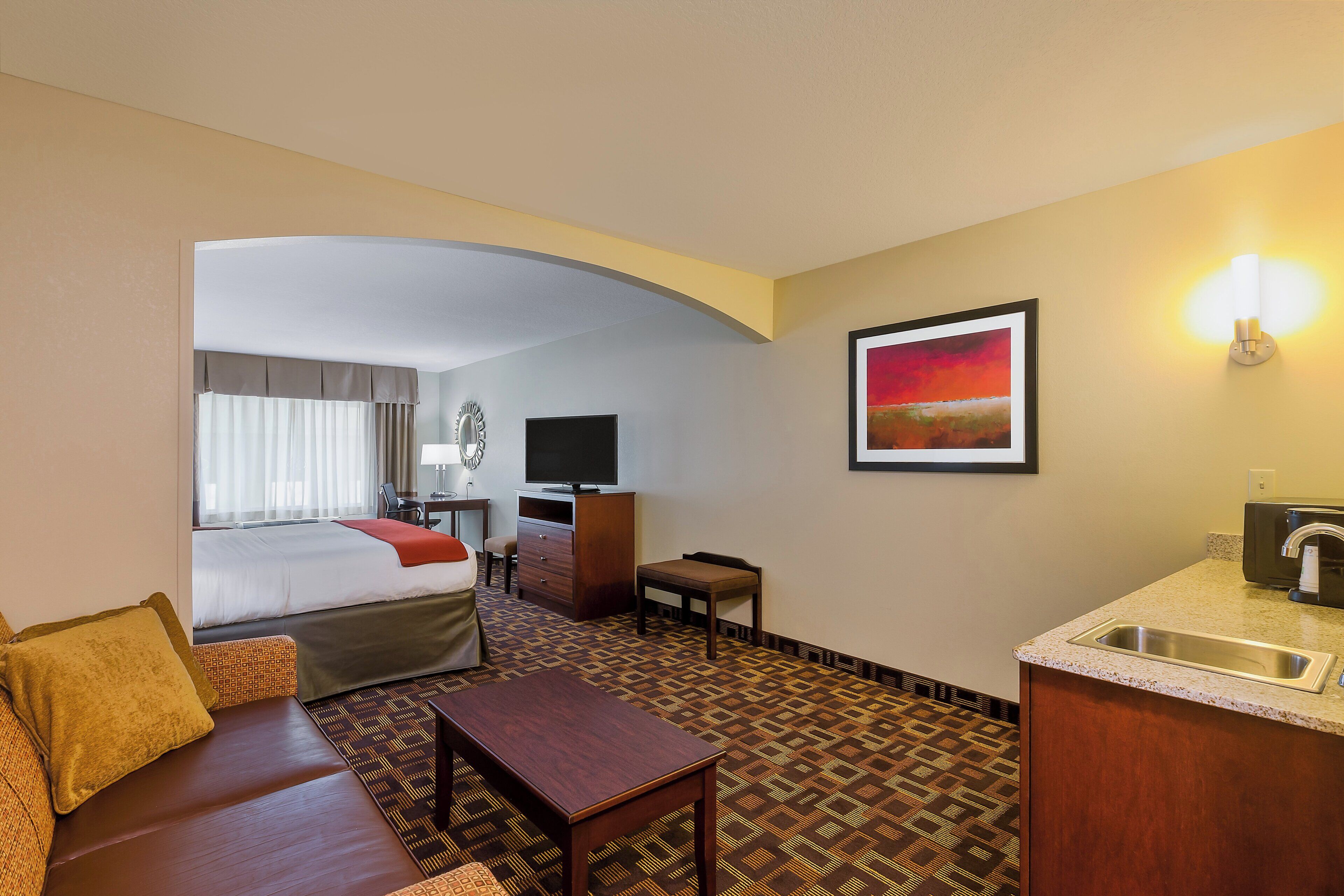 Habitación de hotel con cama, escritorio, sillón, sofá, televisión, cuadro y kitchenette.