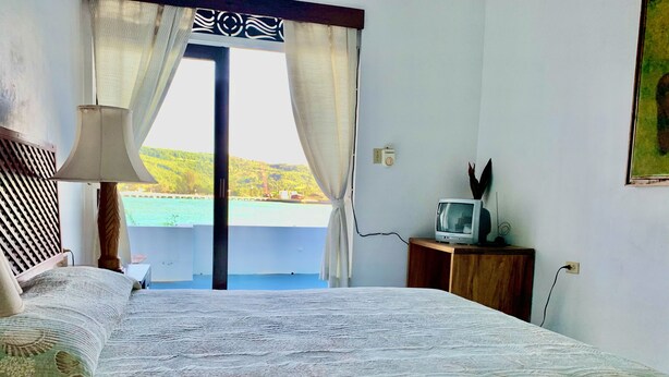 A bedroom with a bed, a TV on a wooden stand, a window with curtains, and a view of a green landscape. - Hotel Rio Bueno (Rio Bueno)