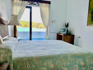 Un dormitorio con cama, televisor sobre un soporte de madera, ventana con cortinas y vistas a un paisaje verde.