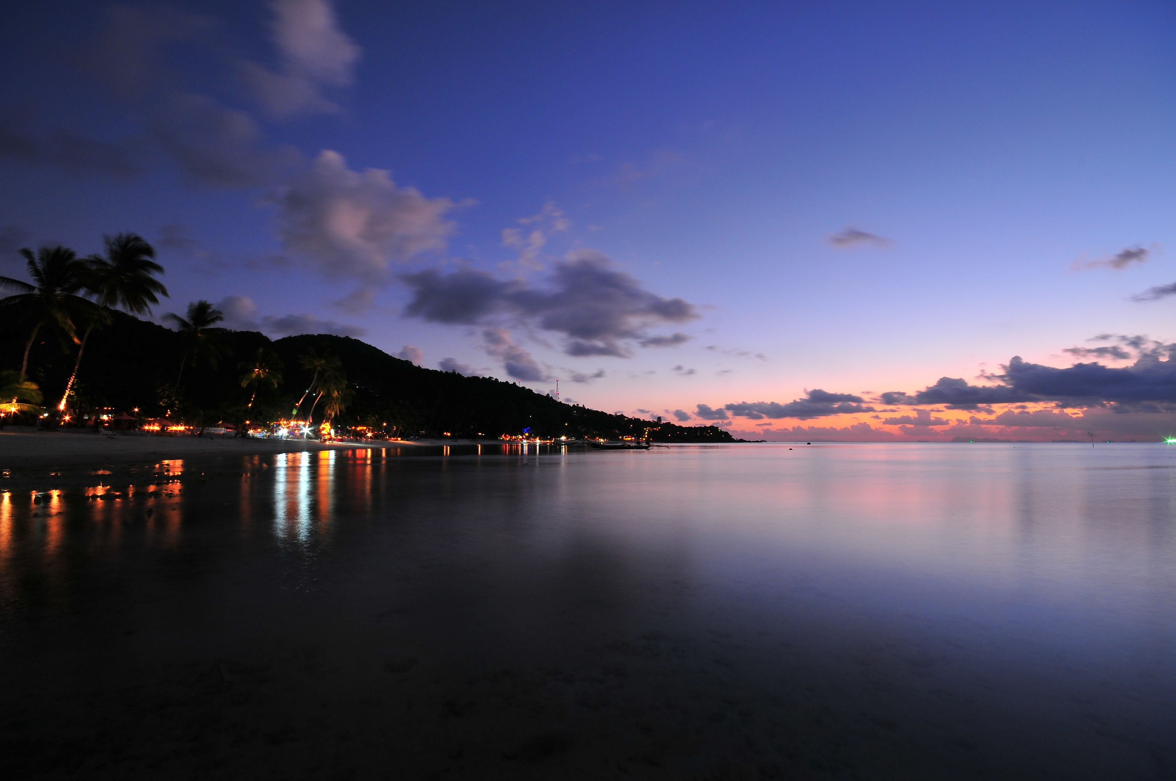 A beach at dusk with calm waters reflecting lights from the shore.