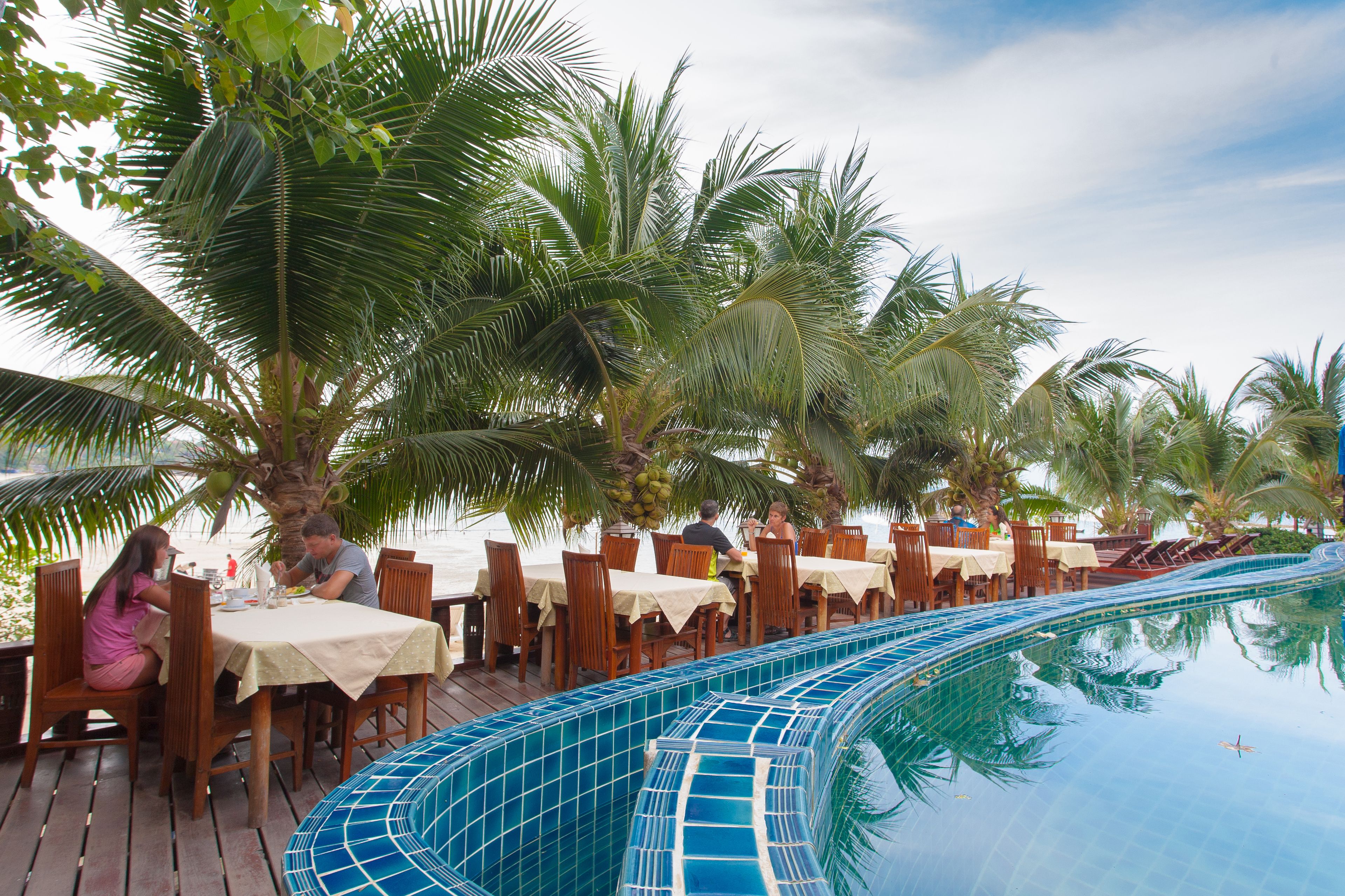 A poolside dining area with wooden tables and chairs, surrounded by palm trees.
