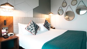 Une chambre d’hôtel comprenant un lit, deux tables de chevet, une petite table, un miroir fixé au mur et un luminaire fixé au mur.