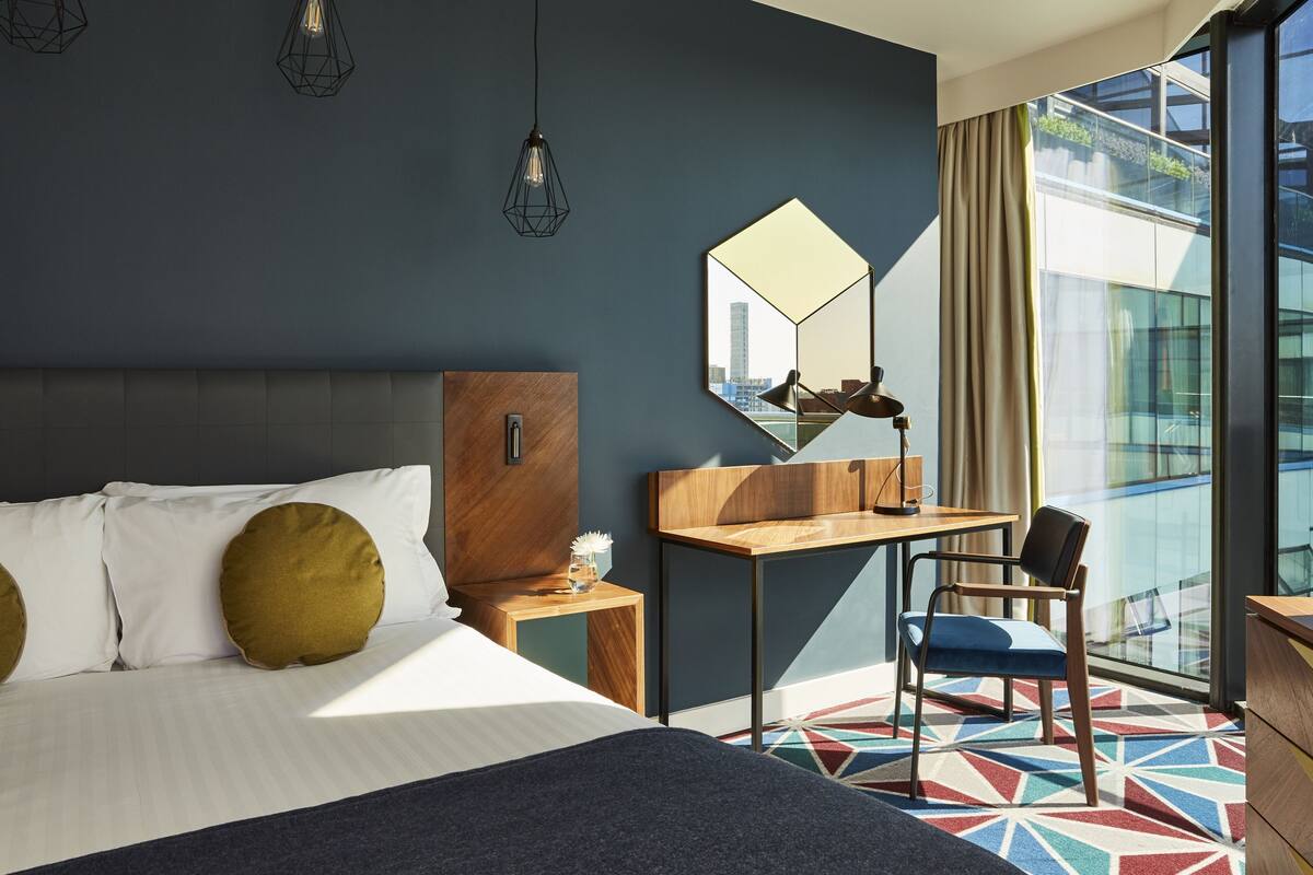 Ein modernes Hotelzimmer mit einem großen Bett, einem Schreibtisch, einem Sessel, einem Spiegel und Blick auf die Stadt durch große Fenster.