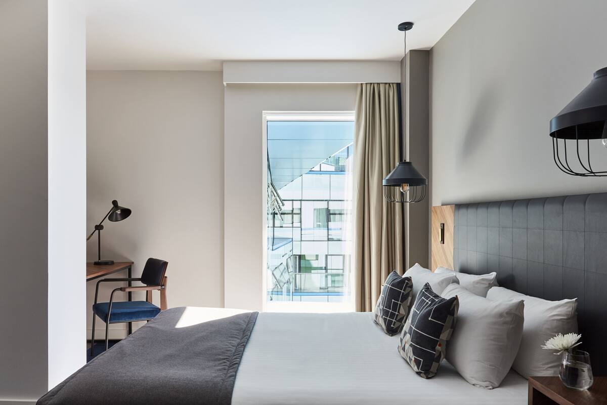 Ein modernes Hotelzimmer mit einem großen Bett, einem Schreibtisch mit Stuhl, einem Fenster mit Blick auf ein Gebäude und einer Lampe auf dem Nachttisch.