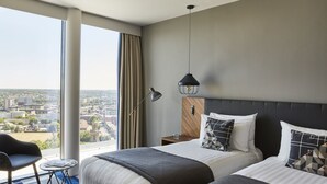 Een hotelkamer met twee bedden, een bureau, een stoel en een groot raam met uitzicht op de stad.