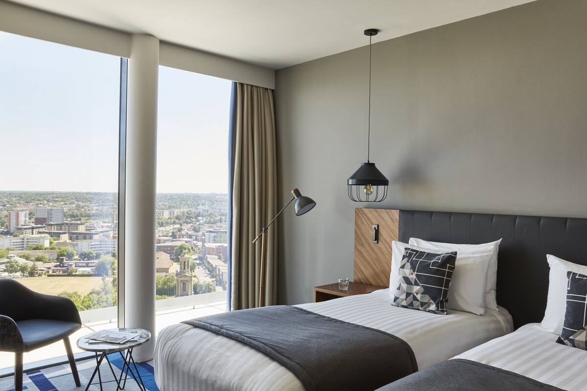 Ein Hotelzimmer mit zwei Betten, einem Schreibtisch, einem Stuhl und einem großen Fenster mit Blick auf die Stadt.