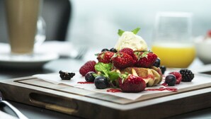 Een ontbijttafel met pannenkoeken belegd met aardbeien, bosbessen en bramen, en een glas jus d'orange op de achtergrond.