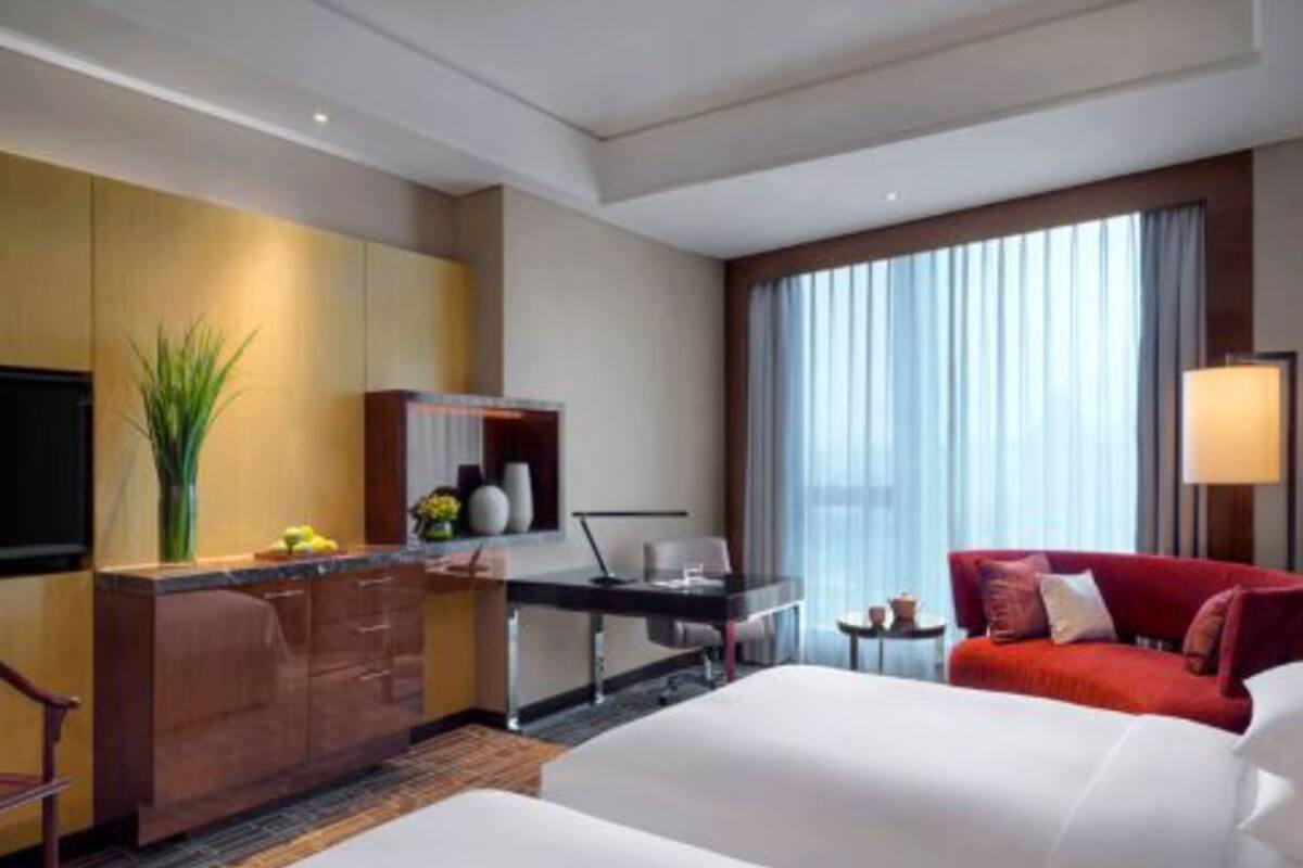 A hotel room with a bed, a desk, a chair, a red sofa, a TV, and a window with curtains.