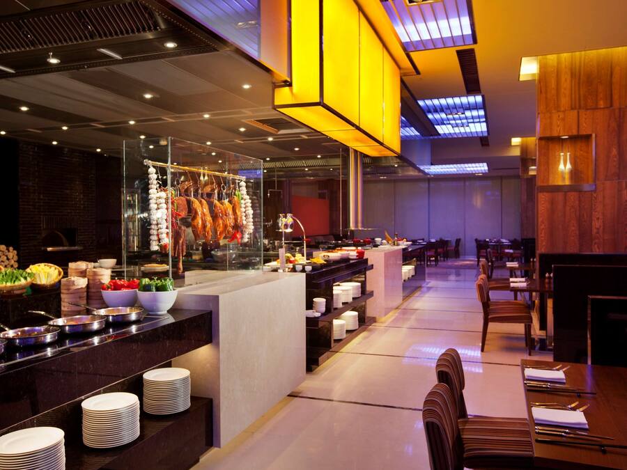 A modern restaurant with a central bar area, multiple dining tables, and a variety of food items displayed on the counter.