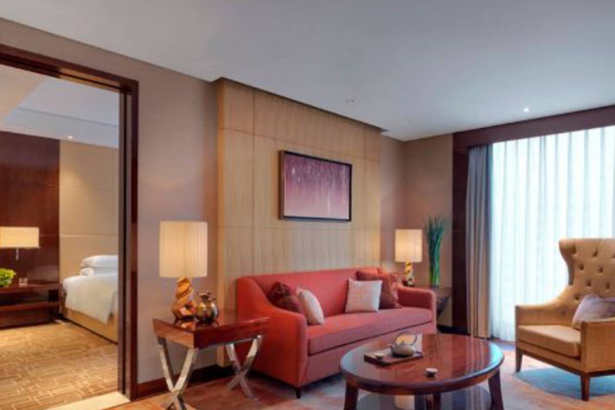 A hotel room with a bed, a red sofa, a wooden coffee table, and a large window with curtains.
