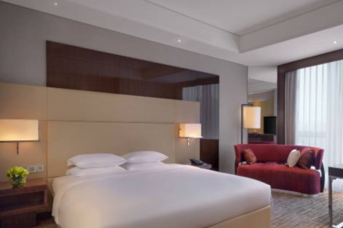 A hotel room with a large bed, two bedside tables, a red sofa, and a large window with curtains.