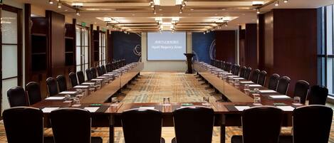 Un salón de conferencias con una larga mesa, sillas, un atril y una pantalla para presentaciones.