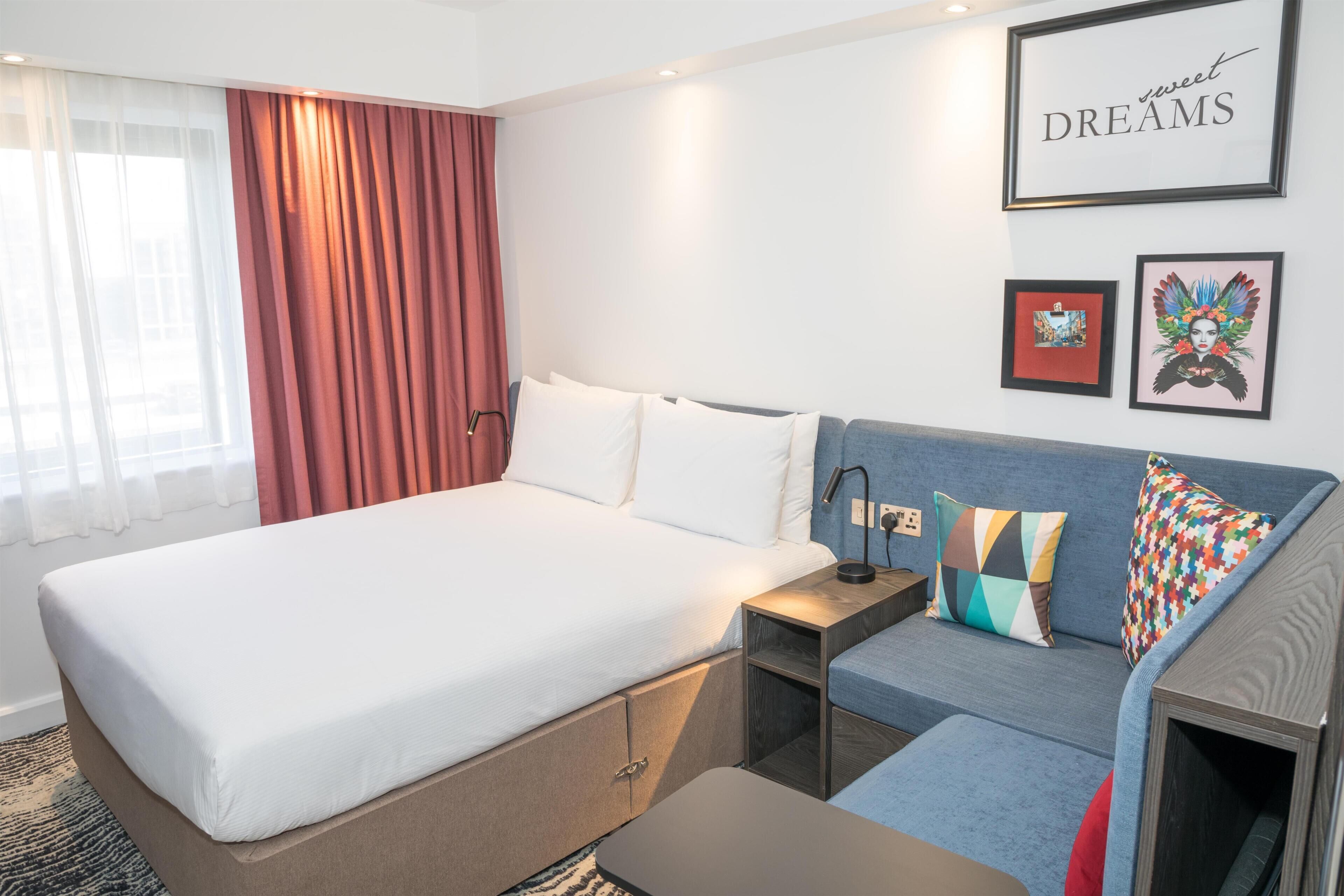 A hotel room with a bed, a sofa, a nightstand, and a wall with framed artwork.