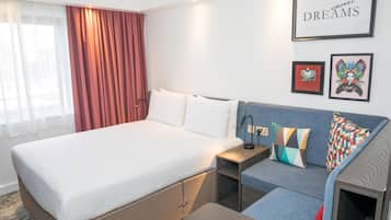A hotel room with a bed, a sofa, a nightstand, and a wall with framed artwork.