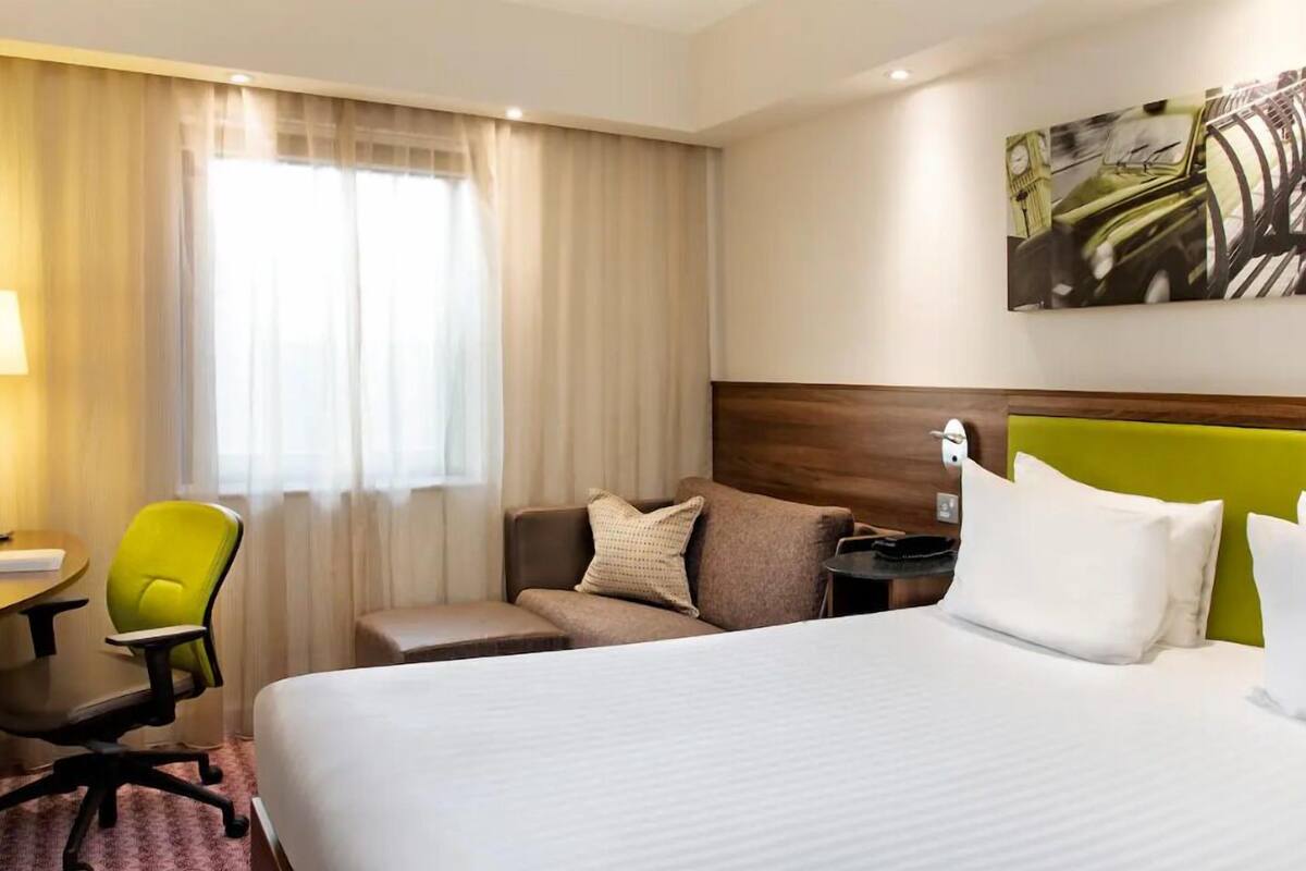A hotel room with a bed, a green armchair, a sofa, a desk, and a painting on the wall.