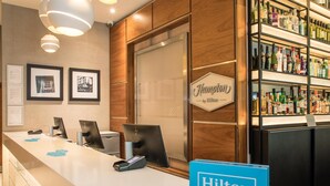 Un espace d’accueil d’hôtel avec un comptoir, des tableaux encadrés accrochés au mur, un panneau indiquant « Hilton Honors » et un présentoir de boissons alcoolisées.
