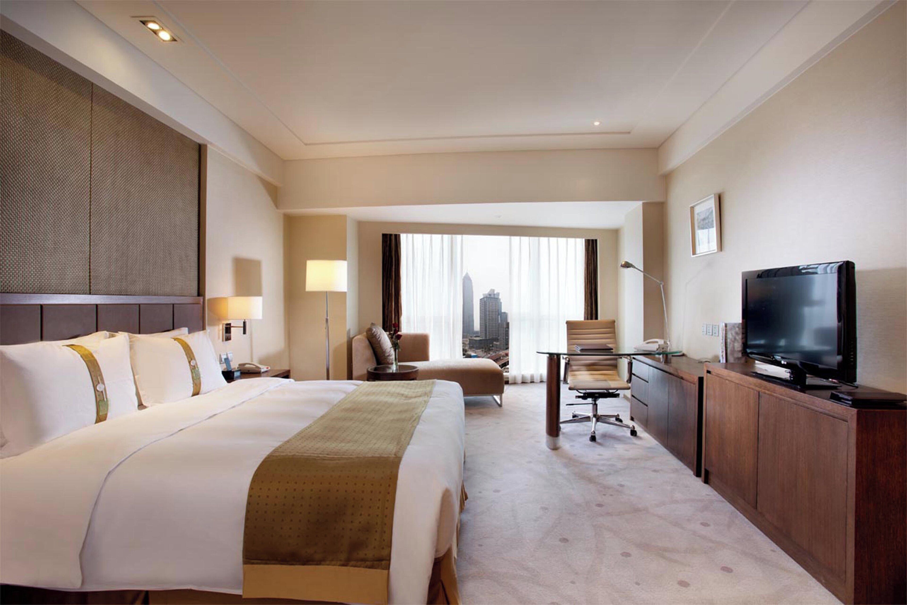 Habitación de hotel con una cama grande, un escritorio, un televisor y vistas de la ciudad a través de la ventana.