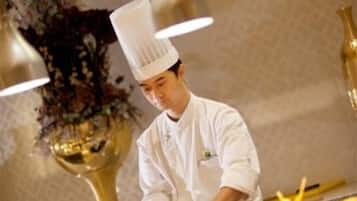 A chef in a white uniform with a tall hat, working at a kitchen counter with a large pot and a decorative vase in the background.