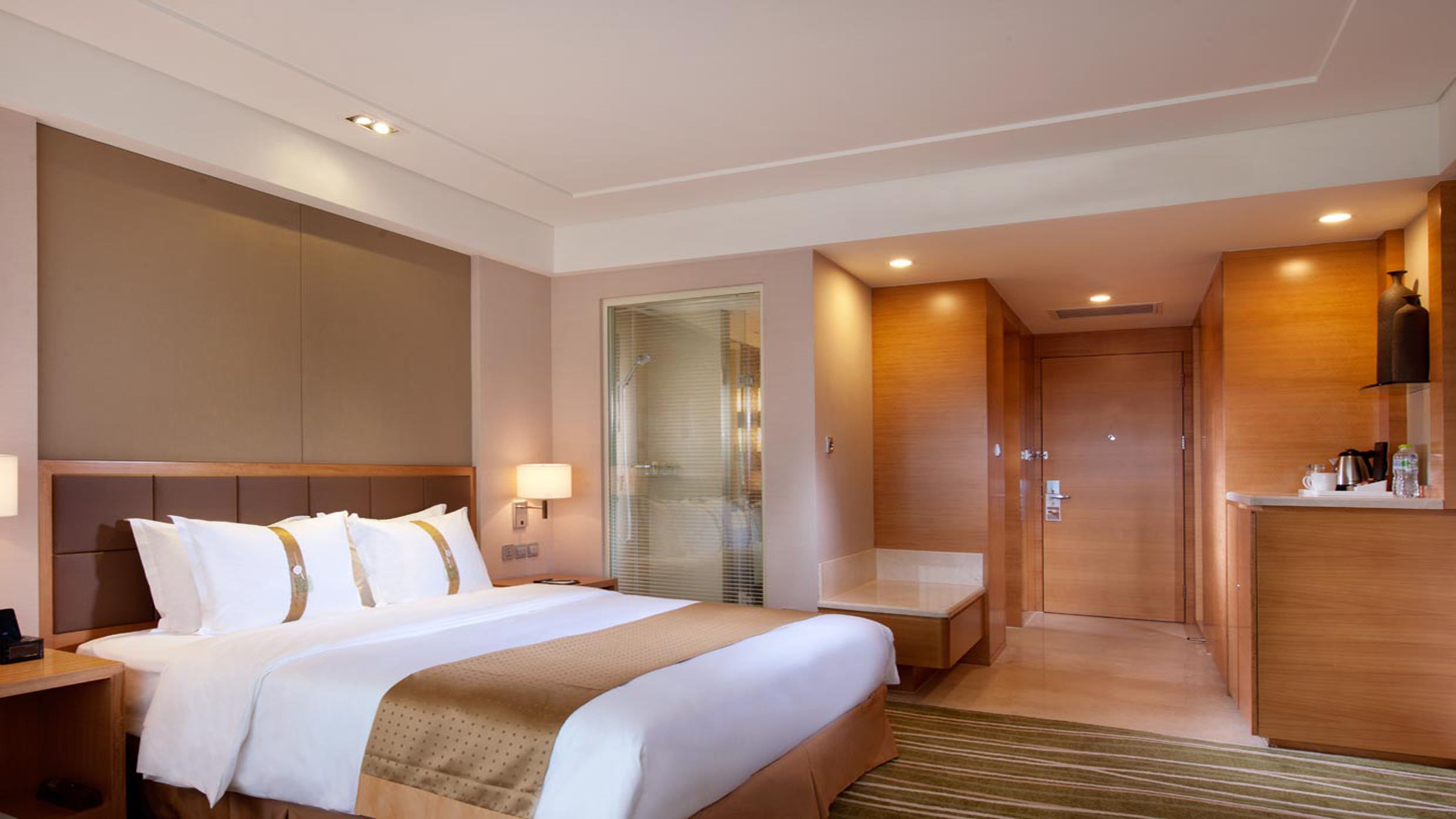 A hotel room with a large bed, a bedside table, a lamp, a shower area, and a kitchenette.