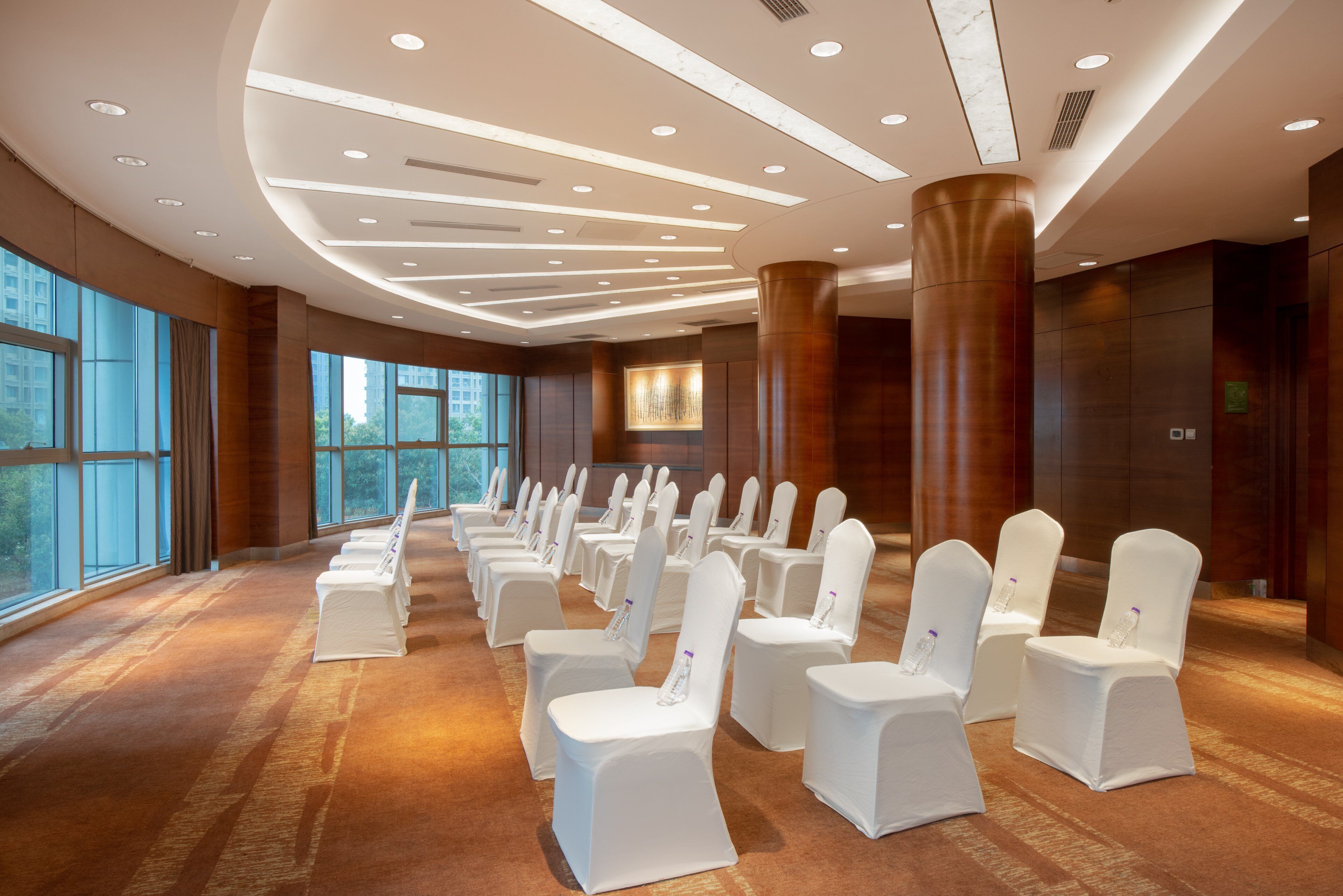 Une salle de conférence avec des rangées de chaises blanches, de grandes fenêtres et un design moderne au niveau du plafond.