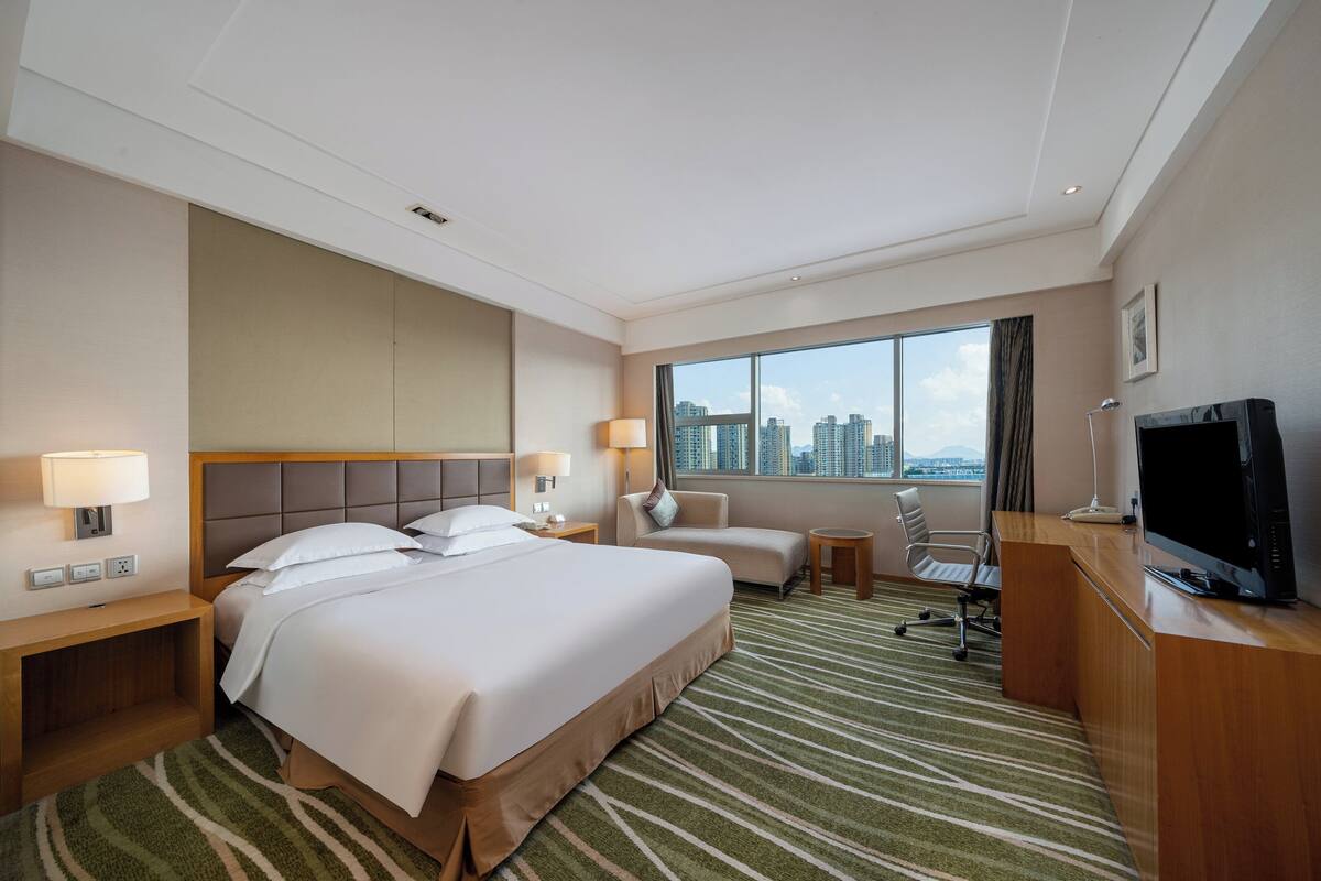 Ein Hotelzimmer mit einem großen Bett, einem Schreibtisch, einem Sessel, einem Fernseher und Blick auf die Stadtlandschaft.