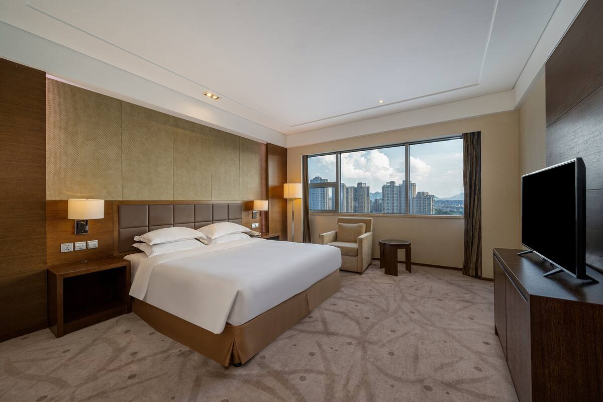 Ein Hotelzimmer mit einem großen Bett, einem Fernseher, einem Sessel und Blick auf die Stadtlandschaft.