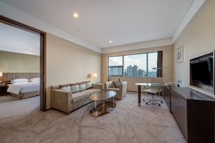 Una habitación de hotel con cama, sofá, mesa de centro, escritorio, televisor y un amplio ventanal con vistas a la ciudad.