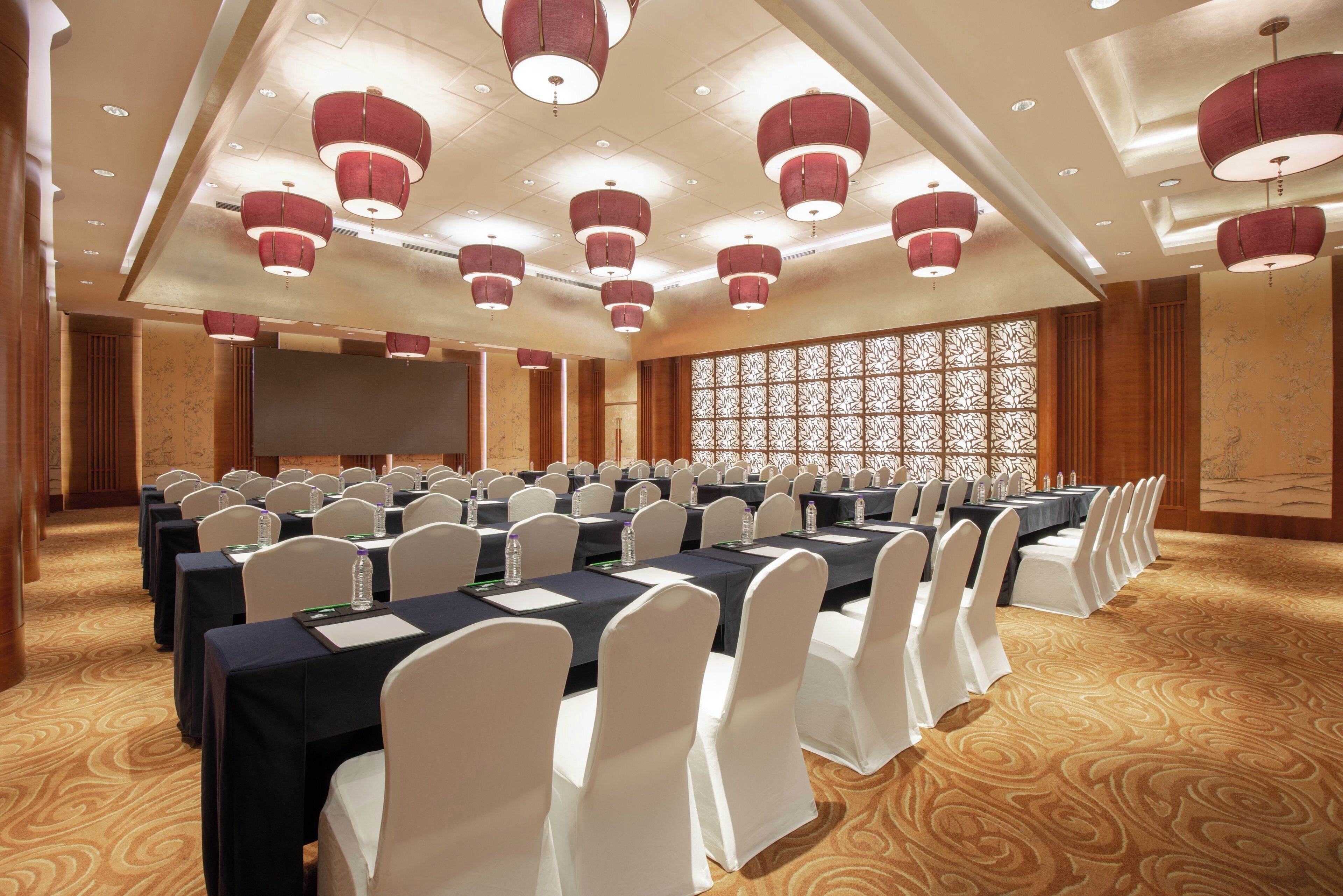 Une salle de conférence avec des rangées de tables et de chaises, un grand écran et un éclairage décoratif.