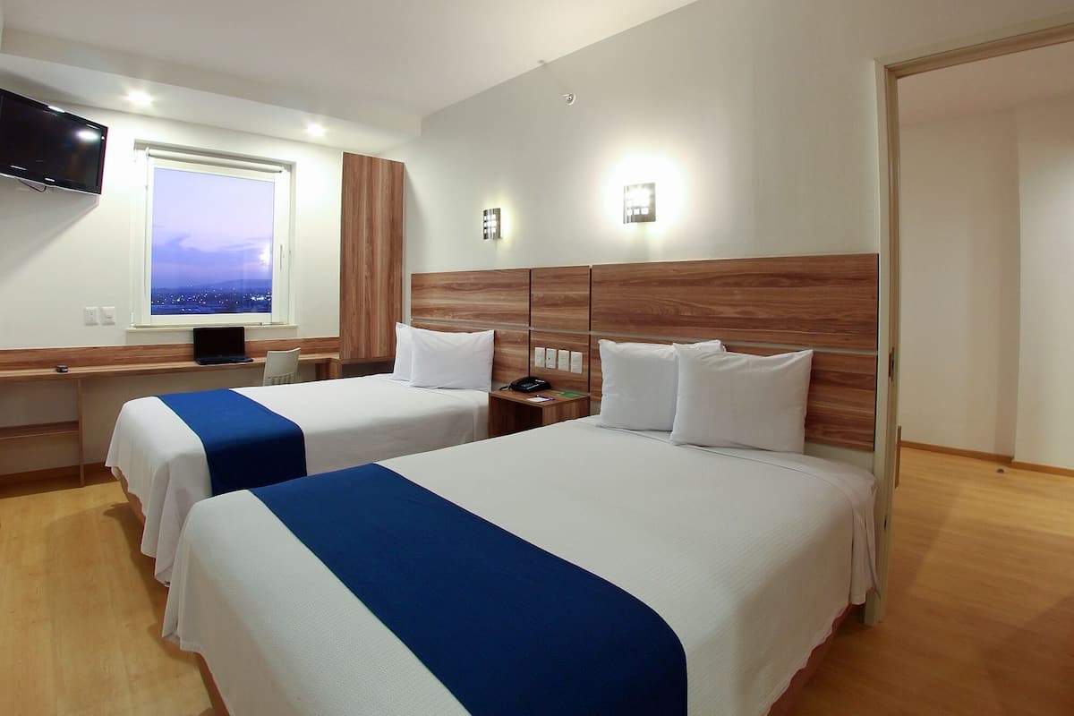 Ein Hotelzimmer mit zwei Betten, einem Fernseher, einem Schreibtisch und einem Fenster mit Blick auf die Stadt.