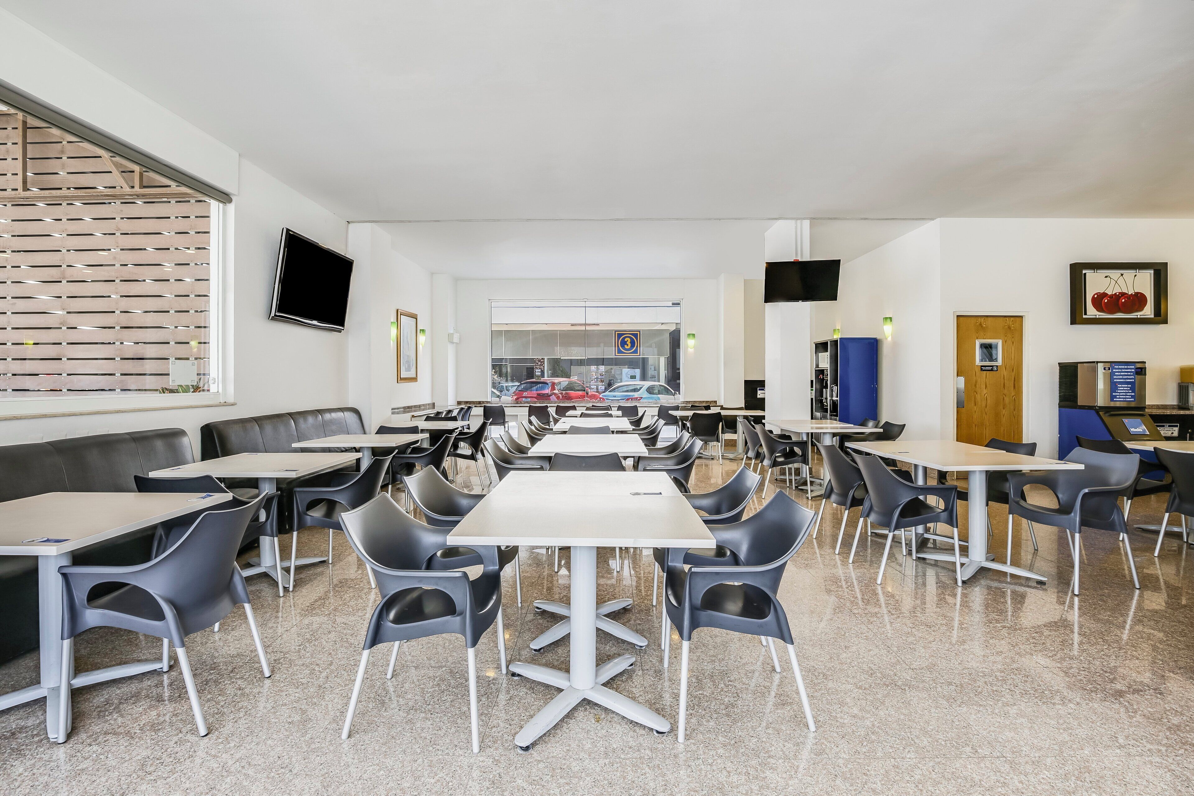 A cafeteria with tables and chairs, a television on the wall, and a window with blinds.