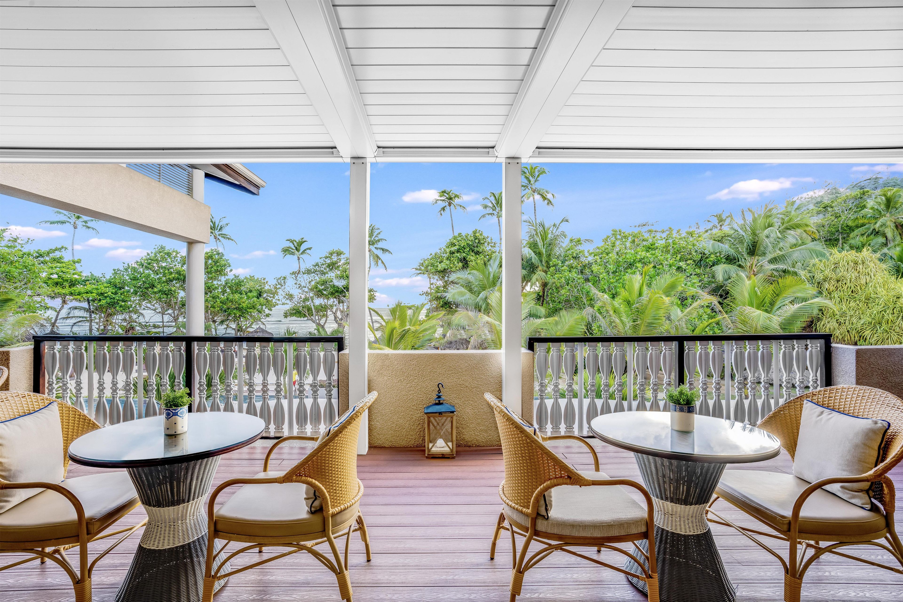 A covered outdoor seating area with wicker chairs and tables, overlooking a lush garden and a clear sky.